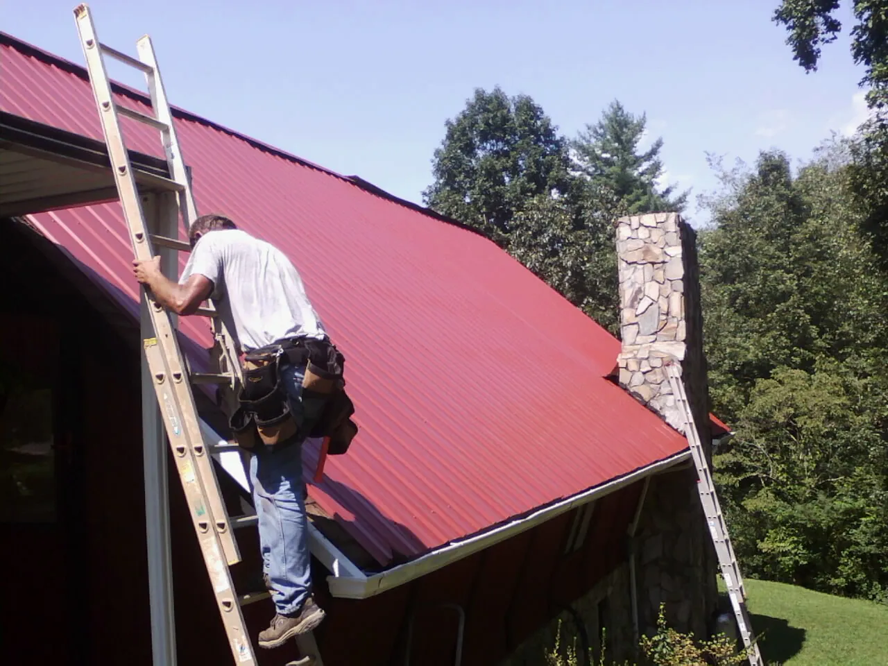 Roofer on a ladder beside a red metal roof and stone chimney — Soffit & Fascia Repair by Roofing Brothers in Bruneau